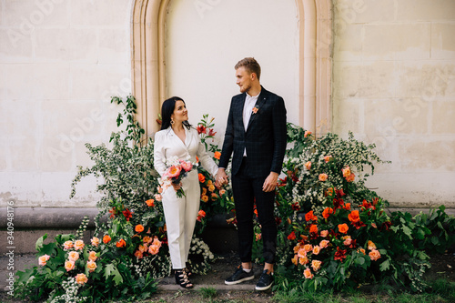 bride and groom posing against the backdrop of an unusual composition of fresh flowers of coral color for an exit wedding ceremony