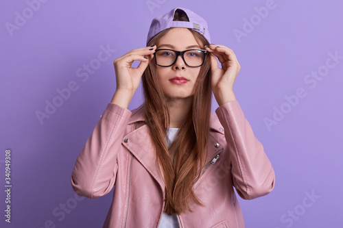 Close up portrait of concentrated serious young female touching eyeglasses frame with fingers, trying new eyewear, looking directly at camera, wearing stylish clothes. Youth and trend concept.