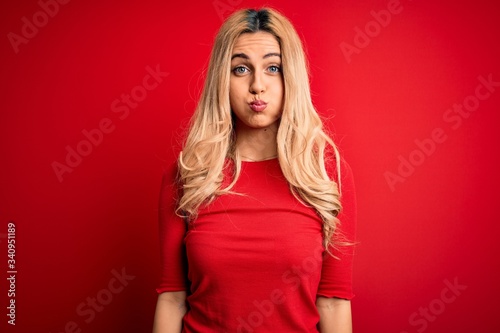 Young beautiful blonde woman wearing casual t-shirt standing over isolated red background puffing cheeks with funny face. Mouth inflated with air, crazy expression.