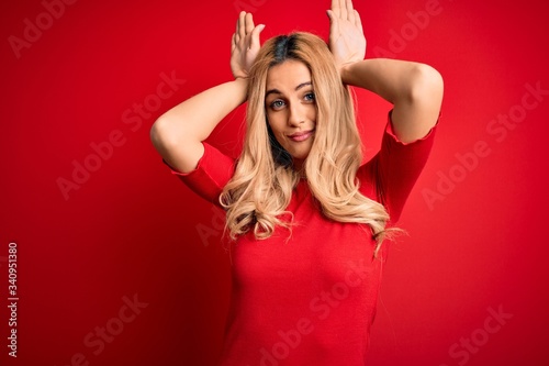 Young beautiful blonde woman wearing casual t-shirt standing over isolated red background Doing bunny ears gesture with hands palms looking cynical and skeptical. Easter rabbit concept.
