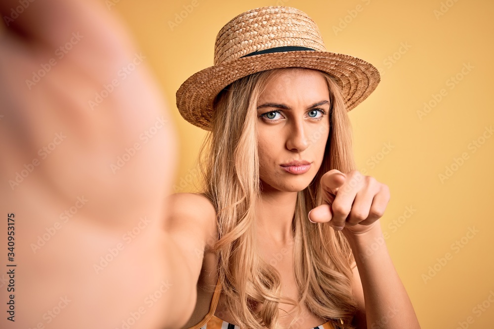 Young beautiful blonde woman on vacation wearing bikini and hat making selfie by camera pointing with finger to the camera and to you, hand sign, positive and confident gesture from the front