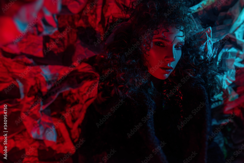 Fashionable beautiful girl with curly hairstyle and makeup. languid sexy look. colorful glare for halloween. newspaper background and black coat