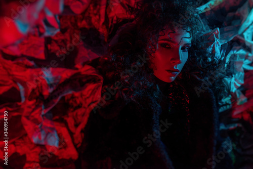 Fashionable beautiful girl with curly hairstyle and makeup. languid sexy look. colorful glare for halloween. newspaper background and black coat