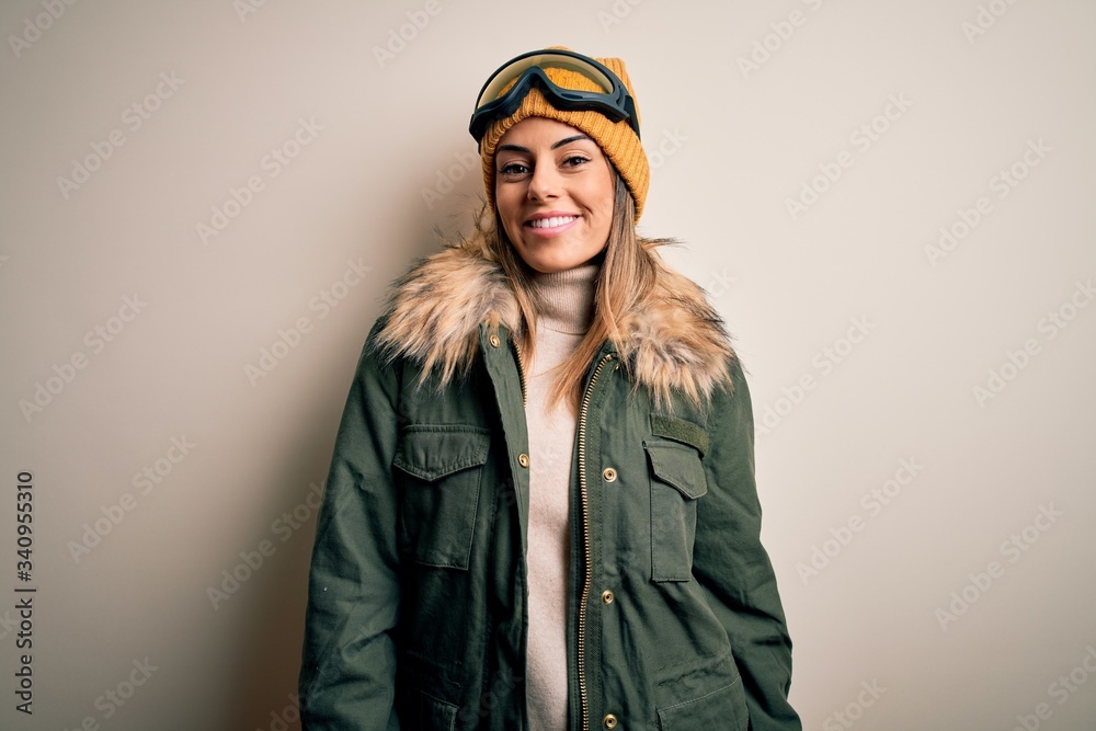 Young brunette skier woman wearing snow clothes and ski goggles over white background with a happy and cool smile on face. Lucky person. Young brunette skier woman wearing snow clothes and ski goggles over white background with a happy and cool smile on face. Lucky person.
