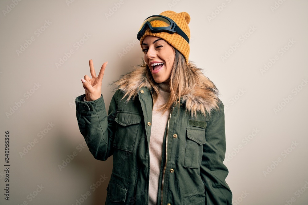 Young brunette skier woman wearing snow clothes and ski goggles over white background smiling with happy face winking at the camera doing victory sign with fingers. Number two. Young brunette skier woman wearing snow clothes and ski goggles over white background smiling with happy face winking at the camera doing victory sign with fingers. Number two.