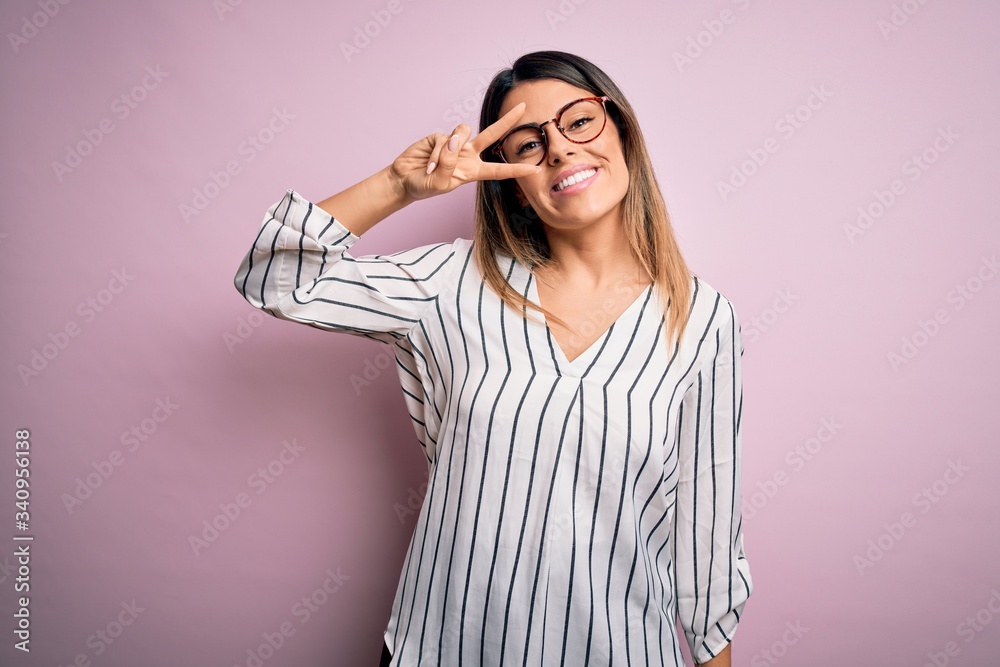 Young beautiful woman wearing casual striped t-shirt and glasses over pink background Doing peace symbol with fingers over face, smiling cheerful showing victory Young beautiful woman wearing casual striped t-shirt and glasses over pink background Doing peace symbol with fingers over face, smiling cheerful showing victory