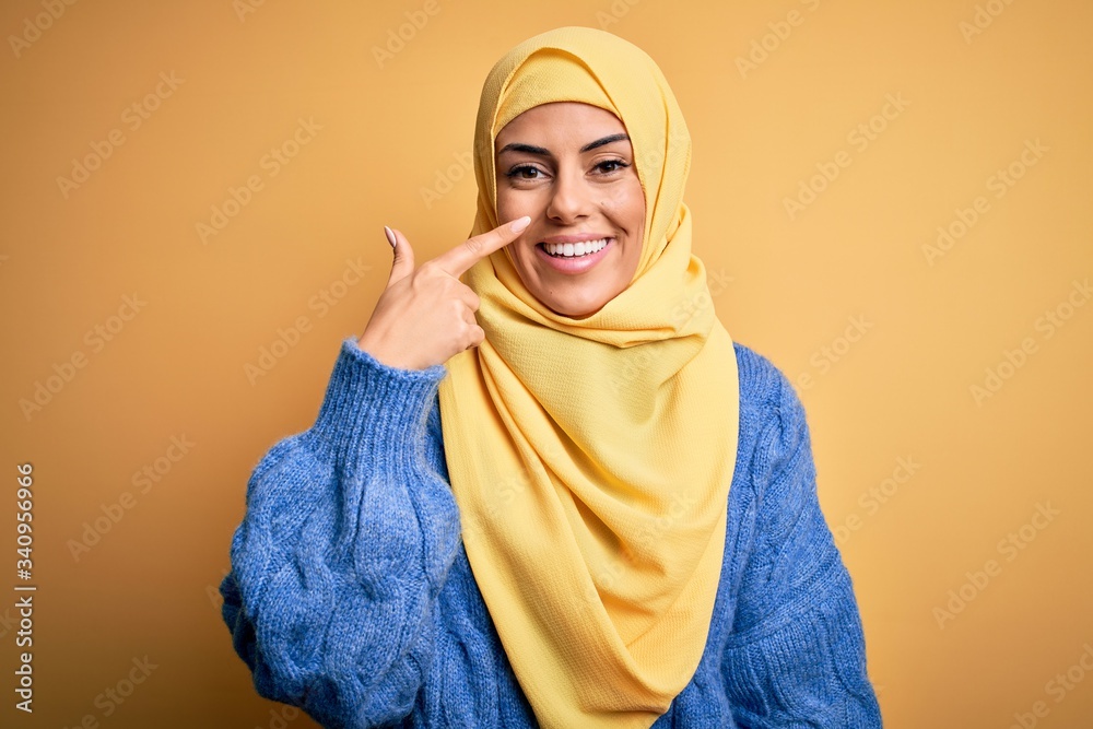 Young beautiful brunette muslim woman wearing arab hijab over isolated yellow background Pointing with hand finger to face and nose, smiling cheerful. Beauty concept