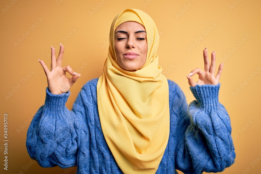 Young beautiful brunette muslim woman wearing arab hijab over isolated yellow background relaxed and smiling with eyes closed doing meditation gesture with fingers. Yoga concept. Young beautiful brunette muslim woman wearing arab hijab over isolated yellow background relaxed and smiling with eyes closed doing meditation gesture with fingers. Yoga concept.