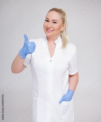 health care and cosmetology concept - portrait of female doctor  nurse or cosmetologist in white uniform and blue gloves thumbs up over gray