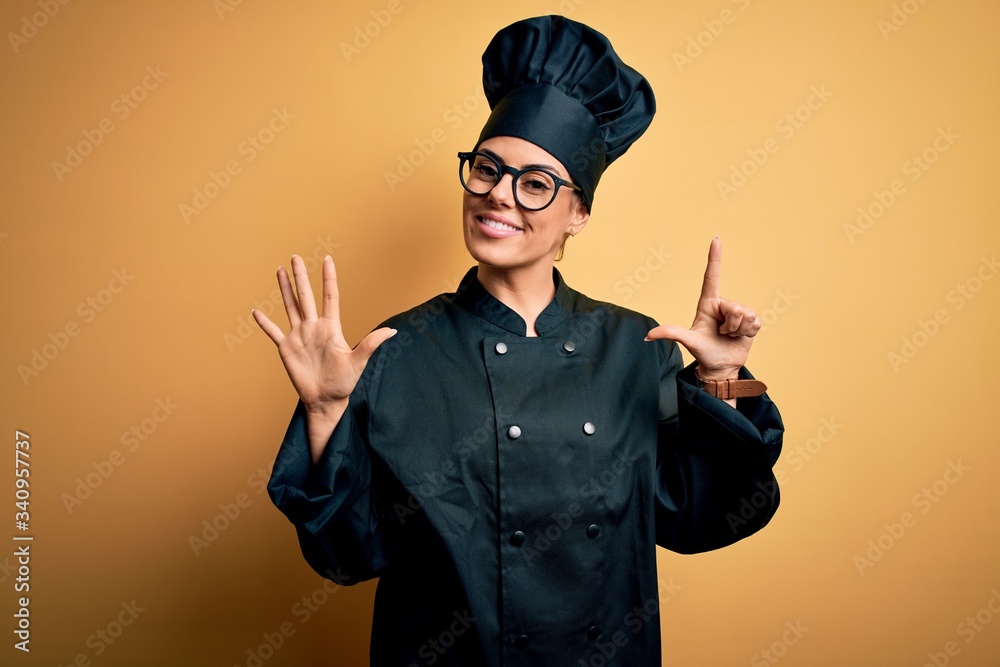 Young beautiful brunette chef woman wearing cooker uniform and hat over yellow background showing and pointing up with fingers number seven while smiling confident and happy. Young beautiful brunette chef woman wearing cooker uniform and hat over yellow background showing and pointing up with fingers number seven while smiling confident and happy.