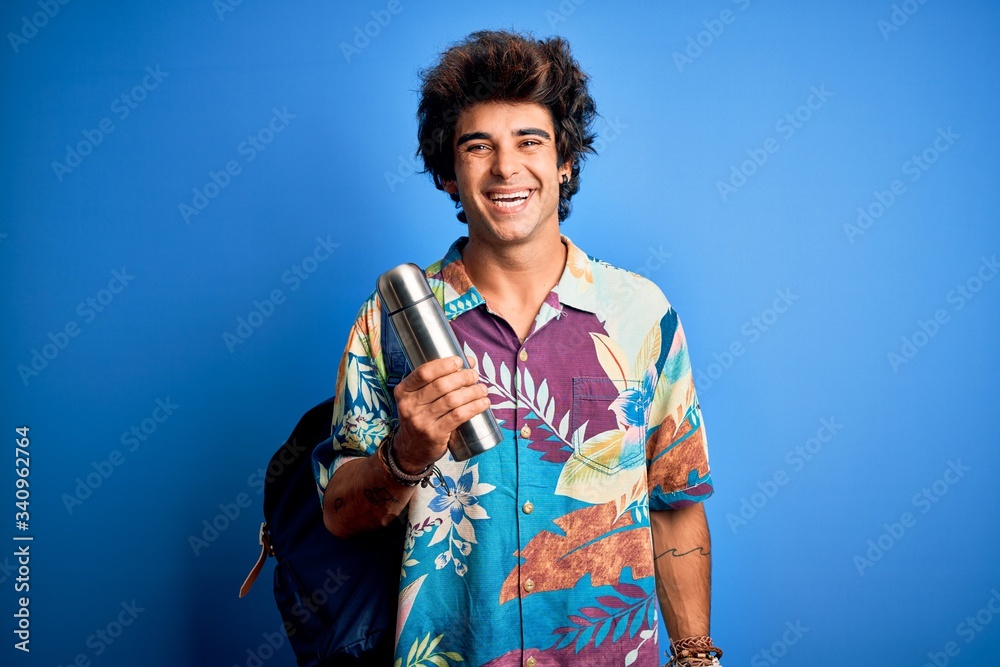 Young tourist man wearing backpack holding thermo over isolated blue background with a happy face standing and smiling with a confident smile showing teeth Young tourist man wearing backpack holding thermo over isolated blue background with a happy face standing and smiling with a confident smile showing teeth