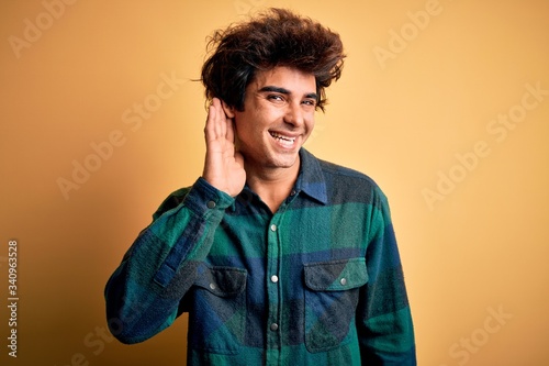 Young handsome man wearing casual shirt standing over isolated yellow background smiling with hand over ear listening an hearing to rumor or gossip. Deafness concept.