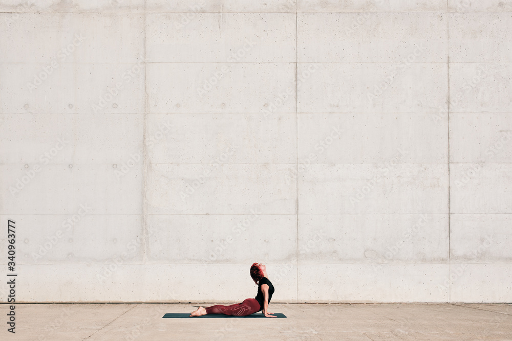 Side view of trendy fit female athlete in sportswear doing baby cobra yoga exercise on sports mat while training alone on street against concrete wall in sunny day Side view of trendy fit female athlete in sportswear doing baby cobra yoga exercise on sports mat while training alone on street against concrete wall in sunny day