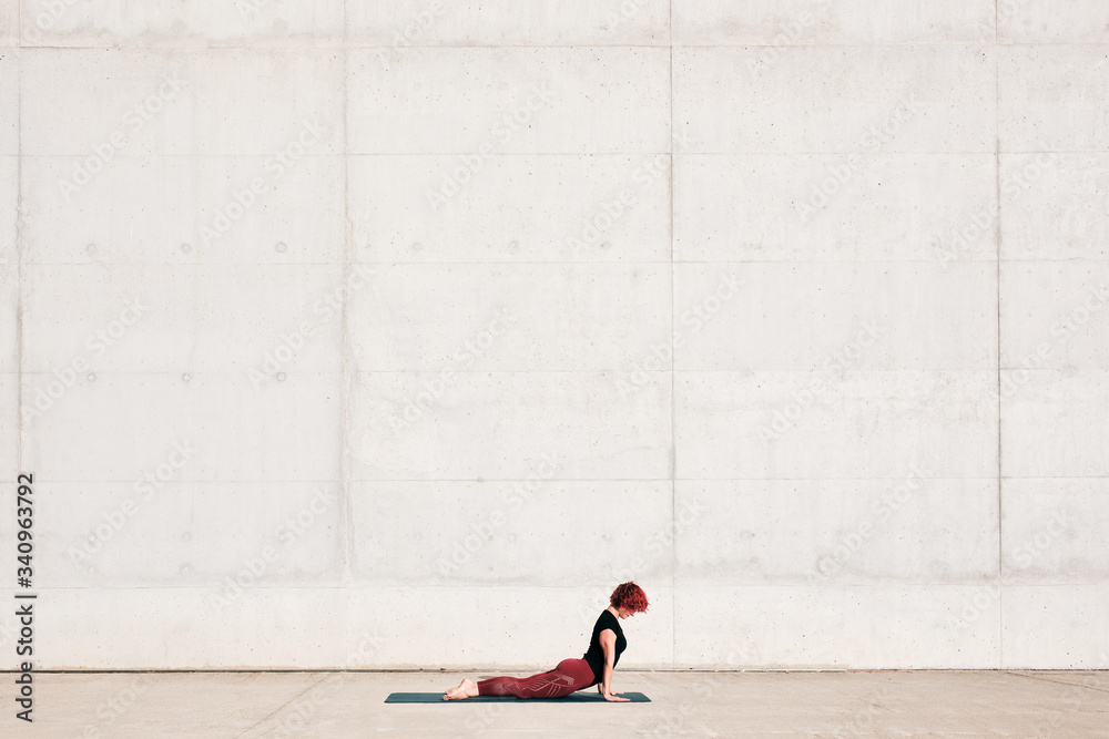 Side view of trendy fit female athlete in sportswear doing baby cobra yoga exercise on sports mat while training alone on street against concrete wall in sunny day Side view of trendy fit female athlete in sportswear doing baby cobra yoga exercise on sports mat while training alone on street against concrete wall in sunny day