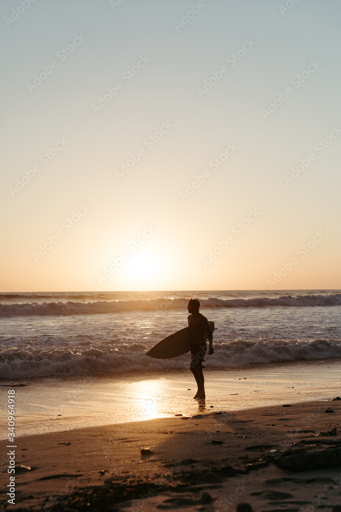 Side view of anonymous man silhouette holding surfboard while walking along sandy seashore in summertime during sunset Side view of anonymous man silhouette holding surfboard while walking along sandy seashore in summertime during sunset