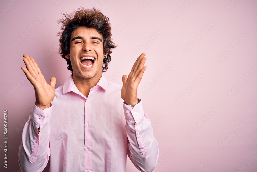 Young handsome man wearing king crown standing over isolated pink background celebrating mad and crazy for success with arms raised and closed eyes screaming excited. Winner concept