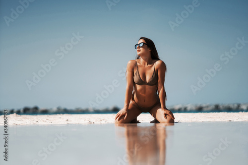 Young sexy woman in sunglasses wearing white swimsuit is posing on the beach