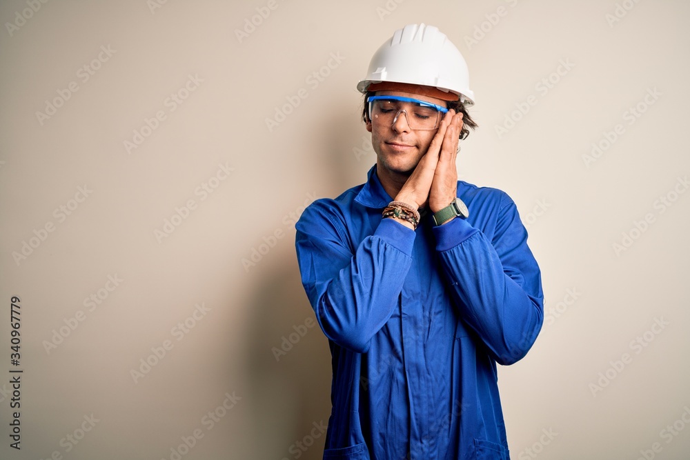 Young constructor man wearing uniform and security helmet over isolated white background sleeping tired dreaming and posing with hands together while smiling with closed eyes.