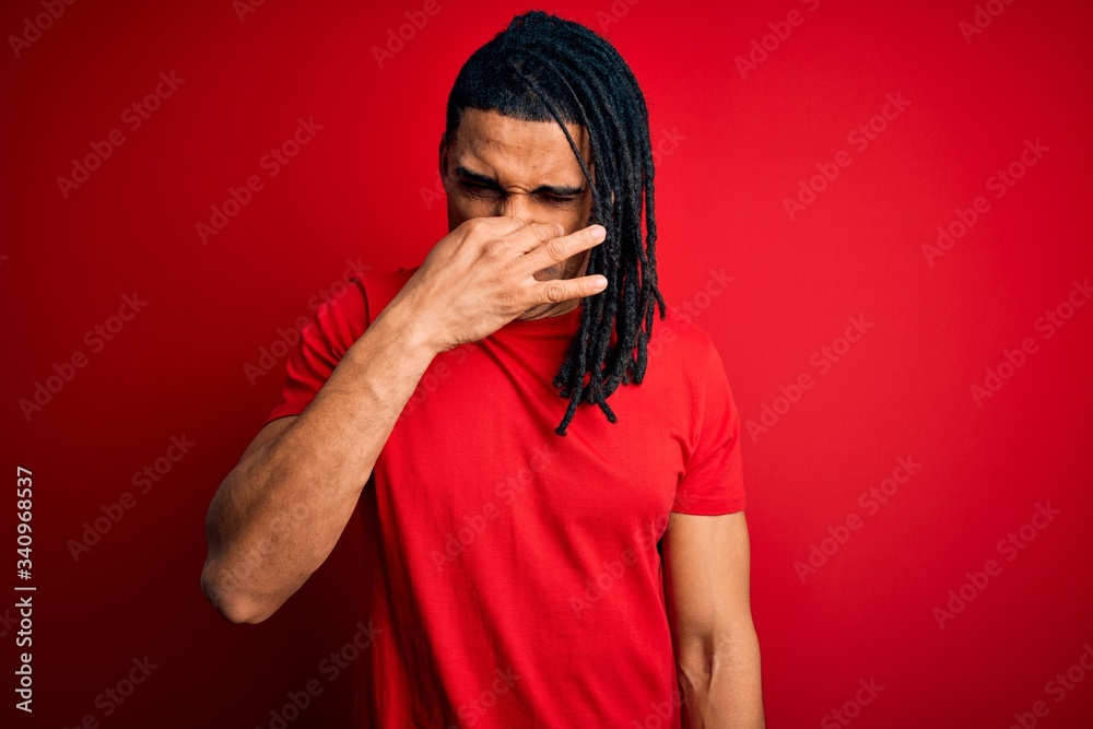 Young handsome african american afro man with dreadlocks wearing red casual t-shirt smelling something stinky and disgusting, intolerable smell, holding breath with fingers on nose. Bad smell