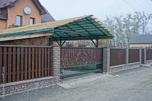 Fototapeta Naklejka Na Ścianę i Meble -  brown metal gate with a black forged pattern and a long fence of wooden boards and bricks on a rural street