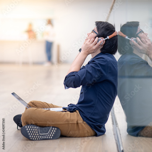 Asian man in casual clothing enjoy listening to music while sitting on the floor with laptop computer and headphones in public building. Urban lifestyle concept