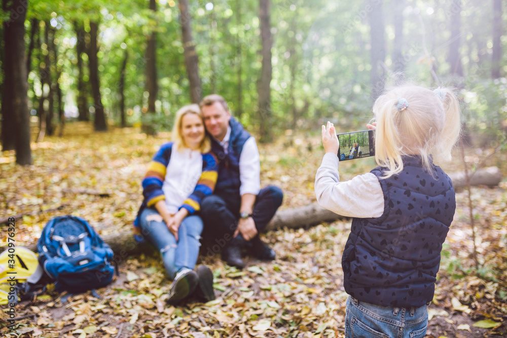 Fototapeta premium travel, tourism, hike, technology and family concept. Happy child makes photo parents in forest. Mom and dad pose for photo, daughter takes photo on phone. Young family of tourists in wooded area