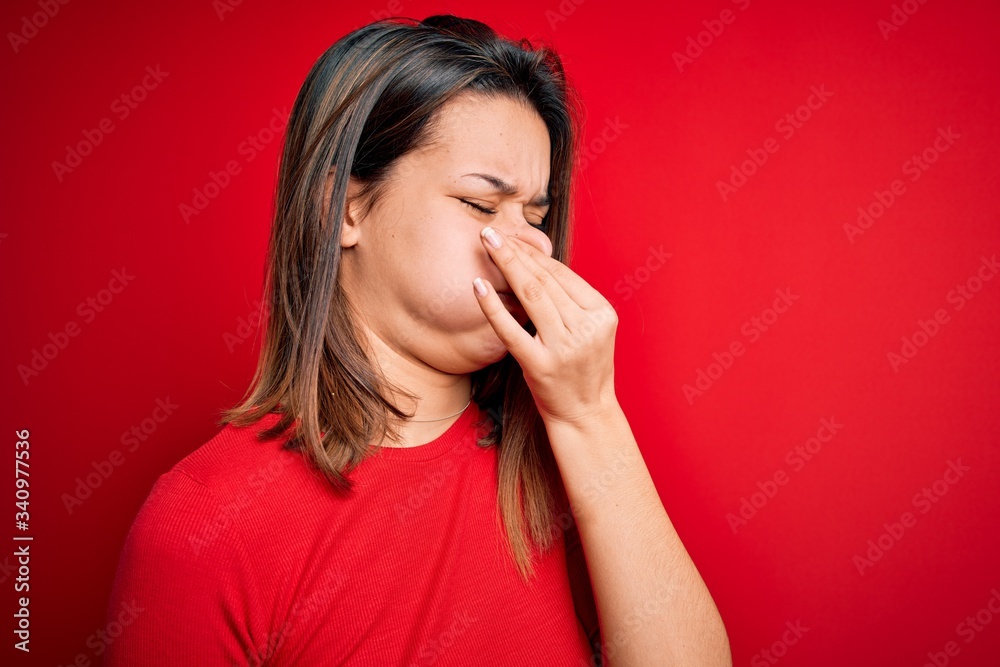 Young beautiful brunette girl wearing casual t-shirt over isolated red background smelling something stinky and disgusting, intolerable smell, holding breath with fingers on nose. Bad smell Young beautiful brunette girl wearing casual t-shirt over isolated red background smelling something stinky and disgusting, intolerable smell, holding breath with fingers on nose. Bad smell