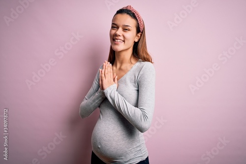 Young beautiful teenager girl pregnant expecting baby over isolated pink background praying with hands together asking for forgiveness smiling confident.