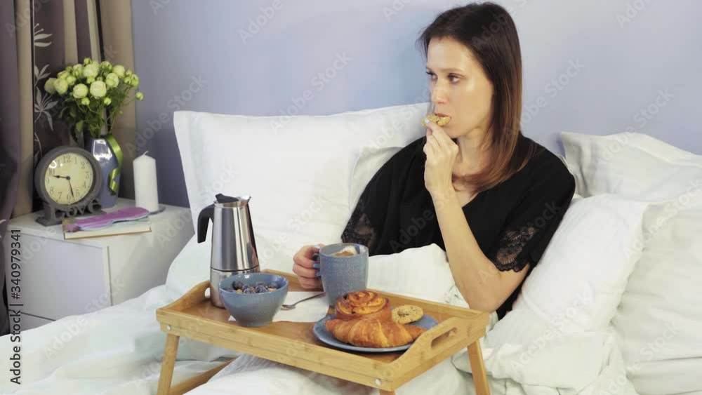 Woman in light hotel apartment lying in bed and have breakfast on small table-tray, enjoying coffee at morning time Woman in light hotel apartment lying in bed and have breakfast on small table-tray, enjoying coffee at morning time