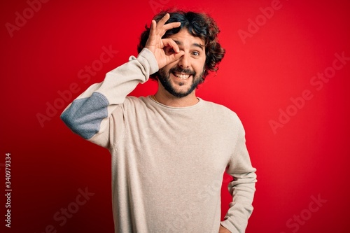 Young handsome man with beard wearing casual sweater standing over red background doing ok gesture with hand smiling, eye looking through fingers with happy face.
