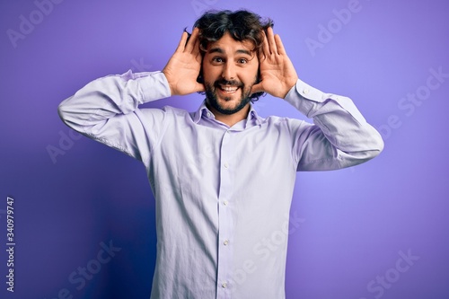 Young handsome business man with beard wearing shirt standing over purple background Smiling cheerful playing peek a boo with hands showing face. Surprised and exited