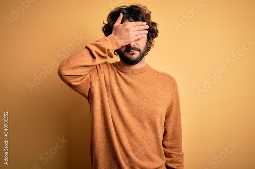 Young handsome man with beard wearing casual sweater standing over yellow background covering eyes with hand, looking serious and sad. Sightless, hiding and rejection concept