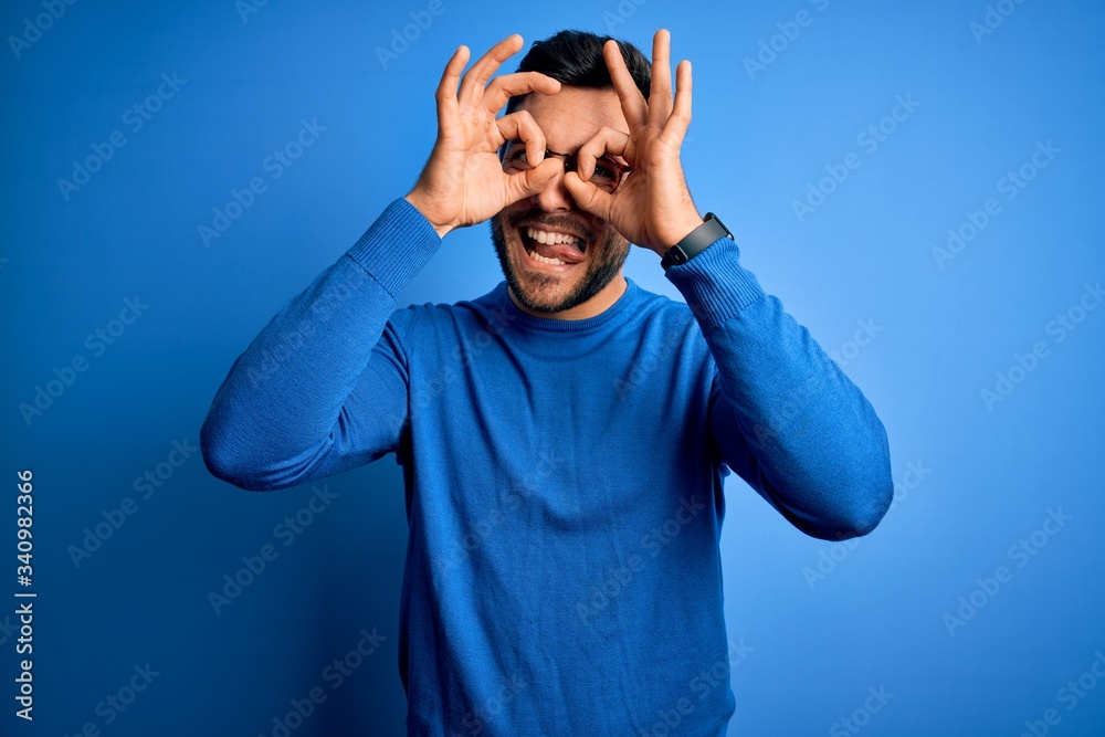 Naklejka premium Young handsome man with beard wearing casual sweater and glasses over blue background doing ok gesture like binoculars sticking tongue out, eyes looking through fingers. Crazy expression.