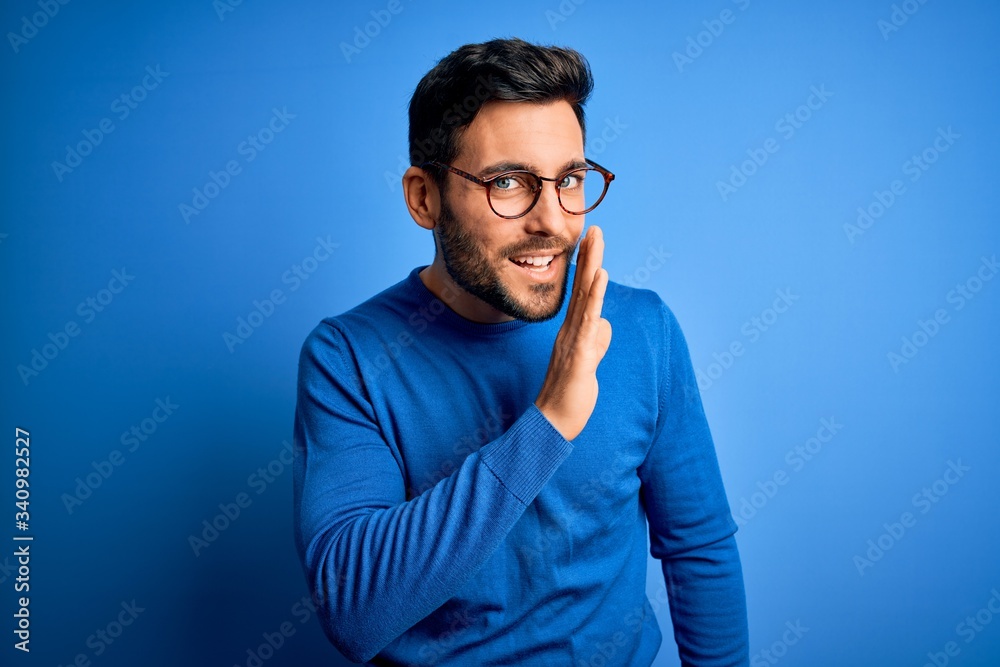 Young handsome man with beard wearing casual sweater and glasses over blue background hand on mouth telling secret rumor, whispering malicious talk conversation - obrazy, fototapety, plakaty 
