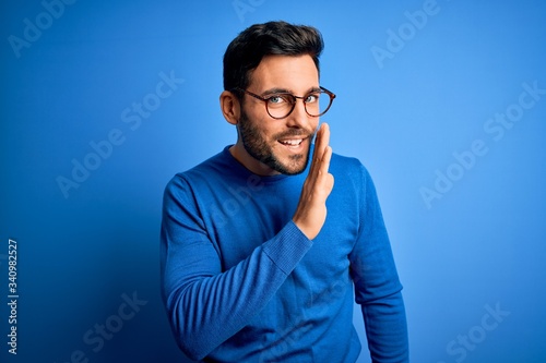 Young handsome man with beard wearing casual sweater and glasses over blue background hand on mouth telling secret rumor, whispering malicious talk conversation photo
