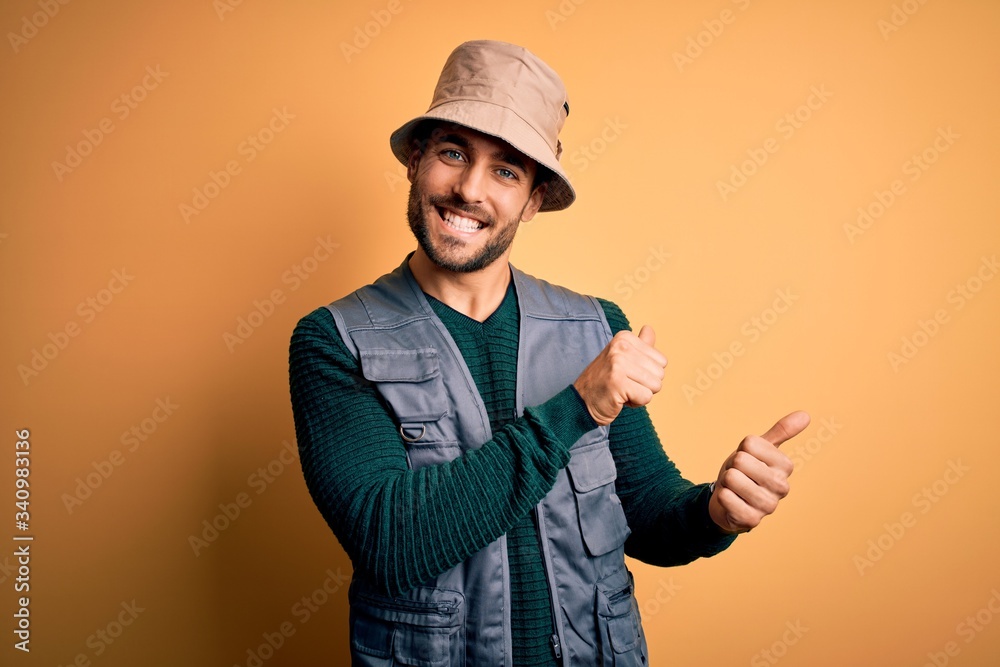 Handsome tourist man with beard on vacation wearing explorer hat over yellow background Pointing to the back behind with hand and thumbs up, smiling confident