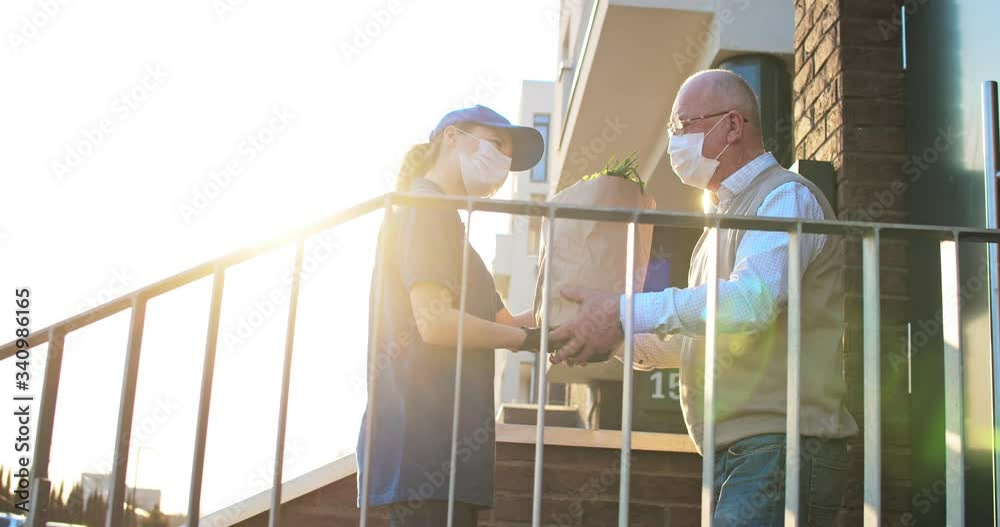 Female courier in medical mask and gloves bringing packet with fresh food to retired old man. Young Caucasian woman handing bag with grocery to male pensioner outdoor at house. Delivery service.