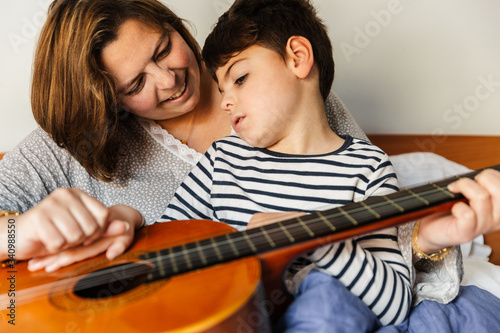 niño juega y aprende con una mujer a tocar la guitarra en la cama. son madre e hijo, él tiene una pluridiscapacidad. Actividad sensorial. están alegres y riendo. instrumento de música. educación.