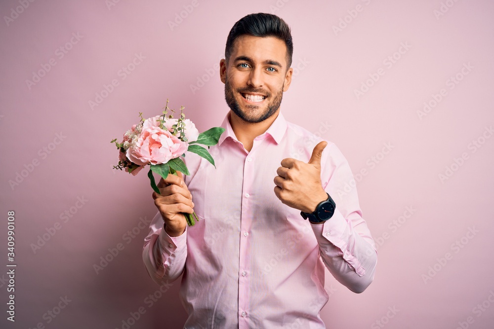 Young romatic man holding bouquet of spring flowers over pink isolatd background happy with big smile doing ok sign, thumb up with fingers, excellent sign Young romatic man holding bouquet of spring flowers over pink isolatd background happy with big smile doing ok sign, thumb up with fingers, excellent sign