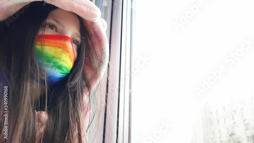Brunette teenager girl in a medical mask painted in bright rainbow colors stands at window with her hand on glass.Concept of staying at home,staying safe.Chasetherainbow flash mob.Distance learning photo