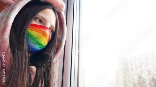 Brunette teenager girl in a medical mask painted in bright rainbow colors stands at window with her hand on glass.Concept of staying at home,staying safe.Chasetherainbow flash mob.Distance learning photo