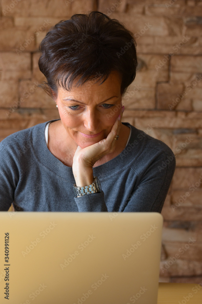 Mature beautiful woman resting chin on hand while using laptop on wooden table against brick wall Mature beautiful woman resting chin on hand while using laptop on wooden table against brick wall