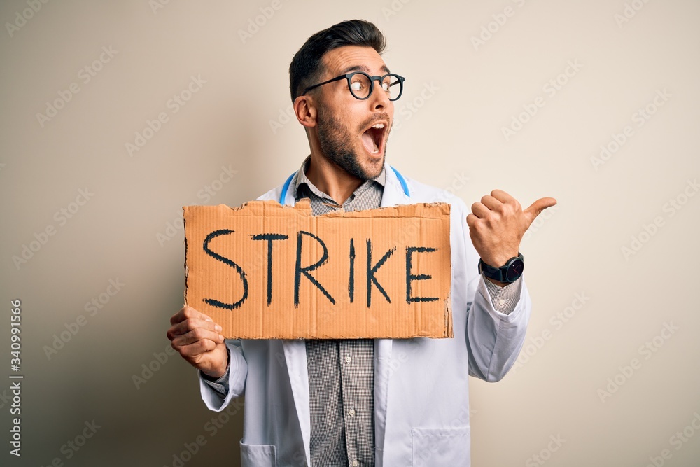 Young handsome doctor man protesting holding cardboard with strike message pointing and showing with thumb up to the side with happy face smiling Young handsome doctor man protesting holding cardboard with strike message pointing and showing with thumb up to the side with happy face smiling