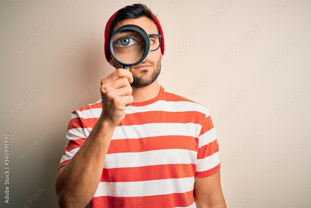 Young detective man looking through magnifying glass over isolated background with a confident expression on smart face thinking serious - obrazy, fototapety, plakaty 