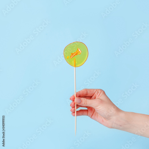 Yellow lollipop in woman hand on blue background. Girls hold hard candy caramel on stick, sweets holidays concept, copy space for text, minimal, summer creative idea - sun and blue sky