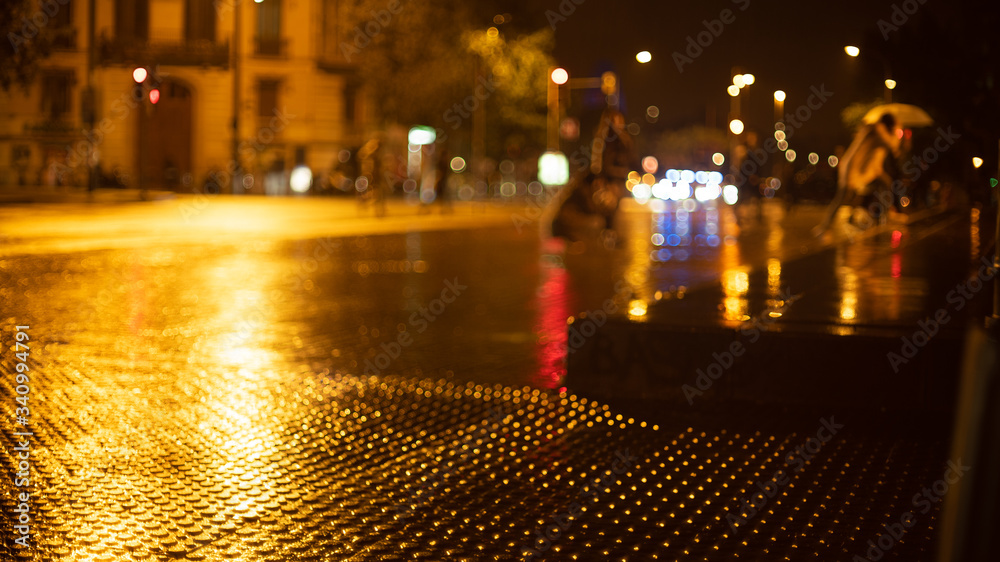 Rainy evening in the city. Blurry figures of people walking. Shiny raindrops on the street. Multi-colored city lights in the background. Creative evening lights and blurred shadows. Twilight mood. 