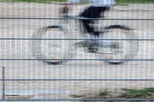 Ein nur teilweise erkennbarer Junge, mit dunkler Hose und blauem Hemd, fährt auf seinem Fahrrad hinter einem Metallgitterzaun. Der Zaun ist scharf abgebildet, der Radfahrer verschwommen und verwackelt