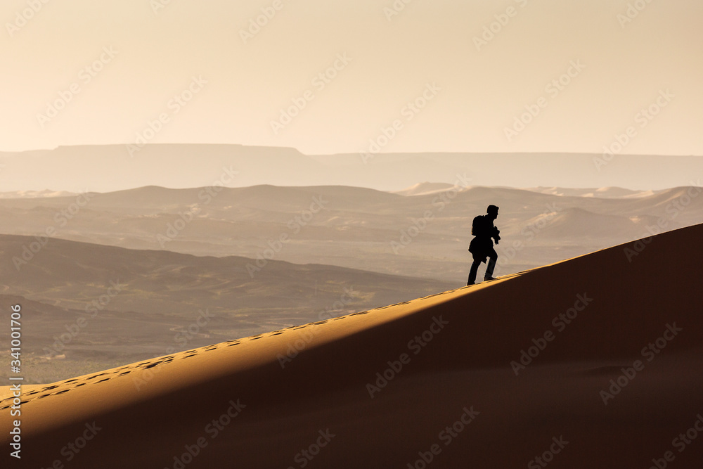 Man walking on dunes of Desert Sahara with beautiful lines and colors at sunrise. Merzouga, Morocco Man walking on dunes of Desert Sahara with beautiful lines and colors at sunrise. Merzouga, Morocco