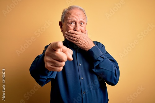 Grey haired senior man wearing casual blue shirt standing over yellow background laughing at you, pointing finger to the camera with hand over mouth, shame expression photo