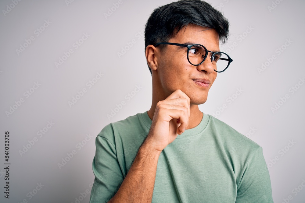 Young handsome man wearing casual t-shirt and glasses over isolated white background with hand on chin thinking about question, pensive expression. Smiling with thoughtful face. Doubt concept. Young handsome man wearing casual t-shirt and glasses over isolated white background with hand on chin thinking about question, pensive expression. Smiling with thoughtful face. Doubt concept.